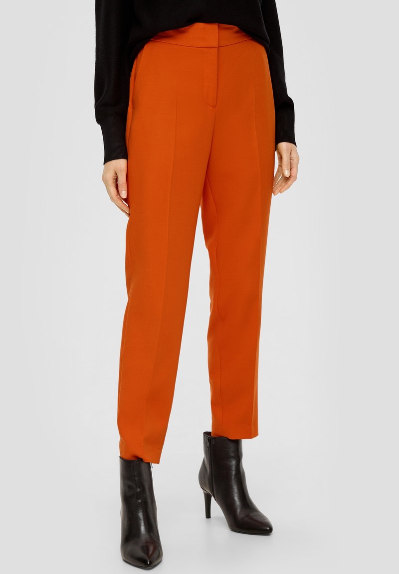 Personne portant un pantalon orange vif ajusté et des bottines noires à talons, debout devant un fond clair uni.