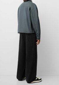 Person som bär en urtvättad blå jeansjacka, vida svarta byxor och svart-vita sneakers, står mot en enkel ljus bakgrund.