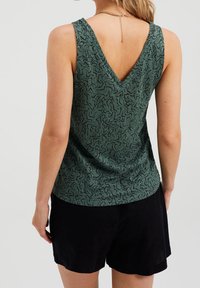 Vihreä hihaton toppi, jossa on V-muotoinen selkäosa, teksturoitu kuvio ja rento istuvuus. Yhdistetty mustiin shortseihin. Kulta ketju kaula-aukossa.