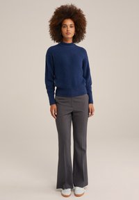 Marinblå, fluffig tröja med hög hals, i kombination med grå utsvängda byxor. Modellen bär vita sneakers, med händerna vid sidorna, på en neutral bakgrund.