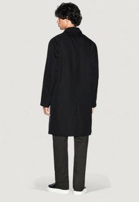 Abrigo negro largo con un diseño limpio, que presenta un corte recto y bolsillos laterales. Lleva puesto sobre pantalones oscuros y zapatillas blancas, mostrando un look minimalista.