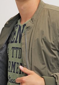 Bunda bomber v olivově zelené barvě, lehká tkanina, ribbované manžety a límec, s předním zipem, přeložená přes zelené grafické tričko.