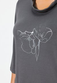 Camiseta gris con un diseño floral plateado, con cuello drapeado y mangas tres cuartos, hecha de una tela suave.