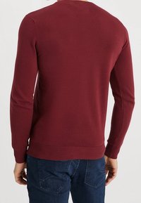 Weinroter Strickpullover mit langen Ärmeln und Rundhalsausschnitt. Glatte Textur mit gerippten Bündchen und Saum. Kombiniert mit blauen Jeans.