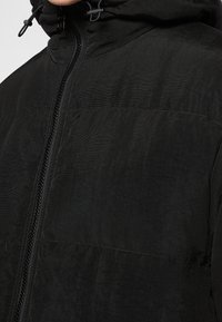 Sort jakke med en struktureret overflade, lynlås foran og justerbar hætte. Har et sømløst design med minimal syning og uden synlige logoer.