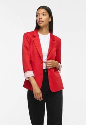 Femme aux longs cheveux foncés portant un blazer rouge sur une chemise blanche et un pantalon noir, regardant vers sa droite avec une main ajustant le blazer.