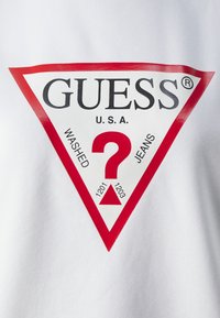T-shirt blanche avec un grand logo triangulaire rouge comportant le texte "GUESS," "WASHED," et "JEANS," ainsi qu'un point d'interrogation rouge au centre.