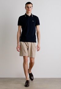 Polo bleu marine avec des accents dorés, shorts beiges et chaussures à enfiler marron foncé. Le mannequin se tient droit contre un fond neutre.