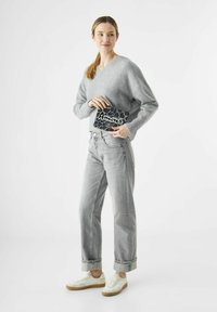 Vrouw in een grijze sweater en jeans houdt een zwarte tas met luipaardprint en de tekst "Amour" vast, terwijl ze tegen een plain witte achtergrond staat.