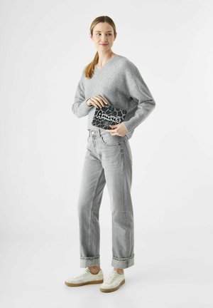 Femme portant un pull gris et un jean, tenant une pochette en imprimé léopard noir avec le texte "Amour", debout contre un fond blanc uni.