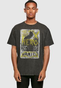 Camiseta negra de gran tamaño con un diseño gráfico de un cartel de Se Busca, con texto y acentos amarillos, que muestra una silueta oscura y tipos de letra en negrita.