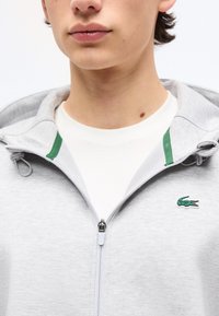 Νέος που φοράει ελαφρύ γκρι φούτερ με φερμουάρ και πράσινο λογότυπο κροκόδειλου στο στήθος πάνω από λευκό πουκάμισο.