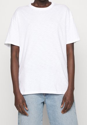 Persoon die een effen wit t-shirt met korte mouwen en lichtblauwe spijkerbroek draagt, staand tegen een neutrale achtergrond.