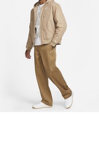 Beige huvjacka, vit grafisk t-shirt, bruna byxor och vita sneakers. Slät textur, avslappnad design, med Nike-logotypdetaljer på jackan och byxorna.