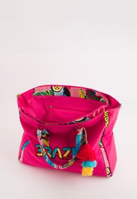 Bolso tote de lona rosa con un interior estampado, texto bordado "BRAZIL" y acentos de borlas coloridas. Presenta una tela y asas resistentes.