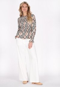 Blusa a maniche lunghe con motivo in nero, pesca e bianco, abbinata a pantaloni bianchi a gamba larga; il modello posa con una mano in tasca.