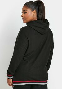 Svart huvtröja i texturerat tyg, med en röd och grå randig fåll. Löst snitt med drop shoulder och en känguruficka.