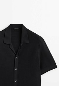 Chemise en maille noire à manches courtes avec un motif texturé, col pointu et fermeture boutonnée sur le devant sur fond blanc.