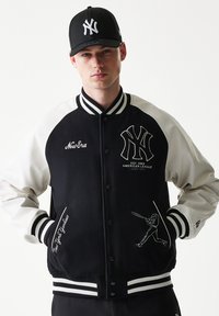 Μαύρο και άσπρο πανωφόρι varsity με λογότυπο των New York Yankees, ριμπ μανσέτες και κεντημένες λεπτομέρειες. Συνδυασμένο με μαύρο καπέλο.
