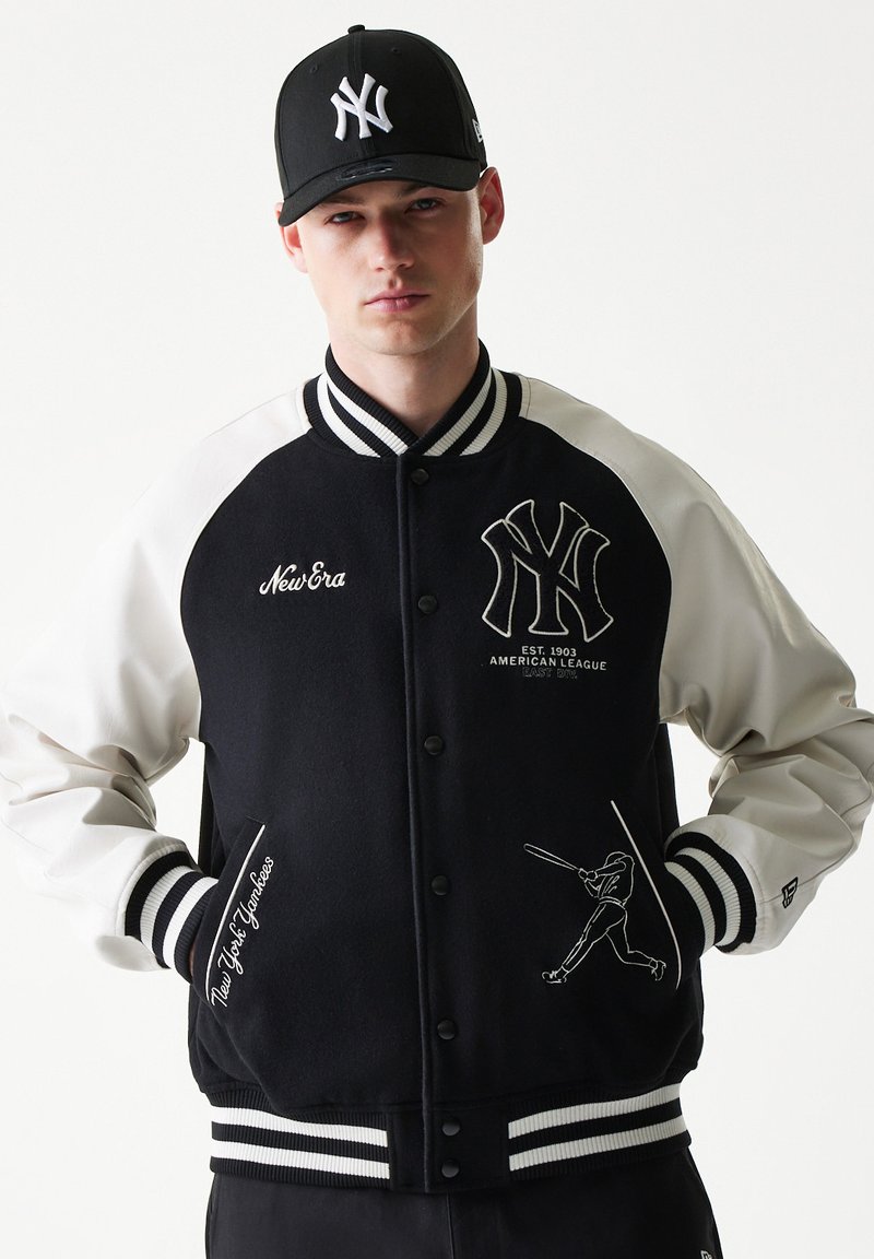 Μαύρο και άσπρο πανωφόρι varsity με λογότυπο των New York Yankees, ριμπ μανσέτες και κεντημένες λεπτομέρειες. Συνδυασμένο με μαύρο καπέλο.