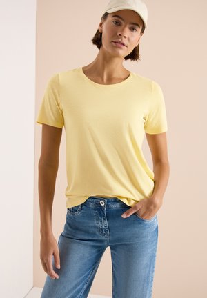 Femme portant une chemise jaune clair à manches courtes, un jean bleu avec une ceinture boutonnée et une casquette de couleur claire, debout devant un fond beige.