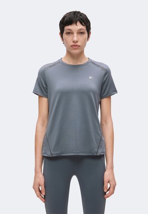 Jong persoon met kort donker haar, gekleed in een aansluitend grijs T-shirt met korte mouwen en bijpassende leggings, staand met het gezicht naar voren en een neutrale uitdrukking.