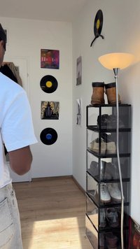 Persoon in witte shirt staat in een kamer met vinylplaten aan de muur, een lamp en een hoge transparante schoenenopslag met verschillende schoenen.