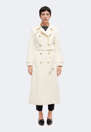 Un mannequin se tient devant un fond uni, vêtu d’un long manteau croisé ivoire avec une ceinture, d’un pantalon noir et de mocassins.