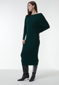 Robe en tricot vert foncé avec une texture côtelée, présentant un col bateau et des manches longues. Associée à des bottes noires à talons pointus.
