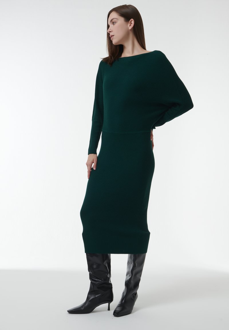 Robe en tricot vert foncé avec une texture côtelée, présentant un col bateau et des manches longues. Associée à des bottes noires à talons pointus.