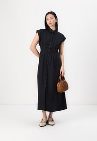 Vestido maxi preto, com botões, mangas curtas e cintura franzida. A modelo segura uma bolsa castanha. Os sapatos são de cor clara e com sola plana.