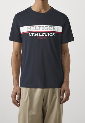 Mężczyzna ubrany w granatowy t-shirt z białym i czerwonym paskiem, napis "HILFIGER ATHLETICS 045/88" oraz beżowe spodnie, stoi na jednolitym tle.