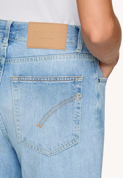 Jeans in denim azzurro chiaro con un'etichetta in pelle beige sulla vita, caratterizzati da cuciture a contrasto e un dettaglio unico con tasca con zip.