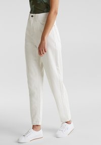 Persona que lleva pantalones de tiro alto de color blanco roto y zapatillas blancas, de pie contra un fondo gris claro liso.