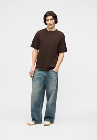 Νέος άνδρας στέκεται με το ένα χέρι στην τσέπη, φορά χαλαρό σκουρό καφέ μπλουζάκι, φαρδιά μπλε τζιν και λευκά αθλητικά παπούτσια, μπροστά σε απλό λευκό φόντο.