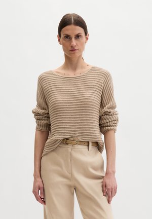 Vrouw met recht, bruin haar die een beige gestreepte gebreide trui, beige hoge taillebroek en een bijpassende riem draagt, staand tegen een witte achtergrond.