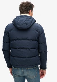Chaqueta acolchada azul marino con capucha, con un tejido texturizado y costuras horizontales. Detalle del logotipo en el hombro. Lleva puesta sobre unos jeans de mezclilla.