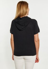 Sweat à capuche noir à manches courtes avec une texture lisse, coupe ajustée et ourlet côtelé, associé à un pantalon de couleur claire.