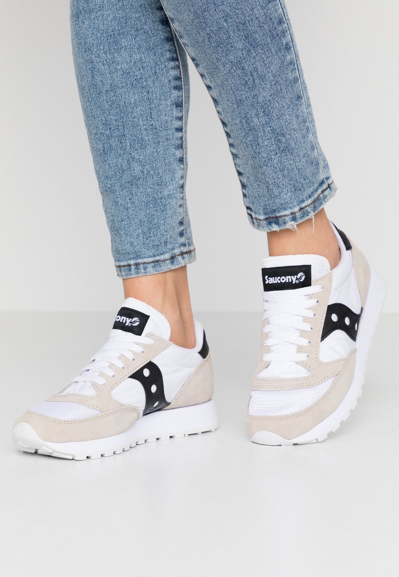 Chaussures de sport Saucony blanches et beige avec logo noir portées avec un jean bleu raccourci sur un fond clair.