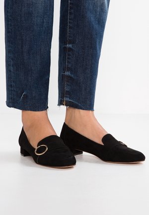 Personne portant un jean court bleu foncé et des chaussures en daim noir à petit talon avec un détail de boucle circulaire dorée.