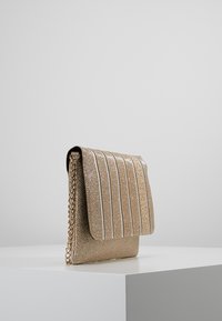 Metaliczna złota torebka o fakturowanym wykończeniu, z pionowymi plisami i łańcuszkowym paskiem. Gładkie zamknięcie na klapę z białymi akcentami.