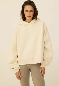 Kremowa, skrócona bluza z kapturem, wykonana z miękkiego materiału, wyróżnia się kapturem, szerokimi rękawami oraz ribbowanym wykończeniem. Połączona z beżowymi legginsami.