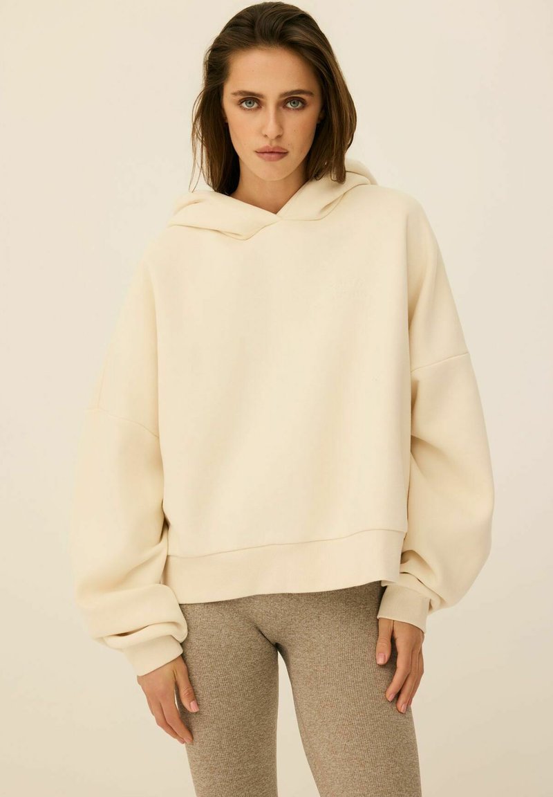 Kremowa, skrócona bluza z kapturem, wykonana z miękkiego materiału, wyróżnia się kapturem, szerokimi rękawami oraz ribbowanym wykończeniem. Połączona z beżowymi legginsami.