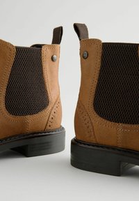 Botas Chelsea de ante marrón con un panel de malla texturizado en marrón oscuro, suela de goma negra y costuras decorativas con lengüetas.