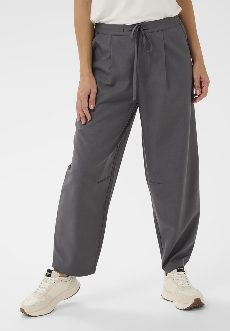 Pantalon gris à jambes larges avec taille à cordon, deux poches latérales et tissu léger. Assorti à des baskets beiges.