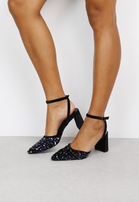 Zapatos de tacón negro con tira en el tobillo que presentan un tacón cuadrado, punta afilada y un upper cubierto de lentejuelas con acentos iridiscentes.