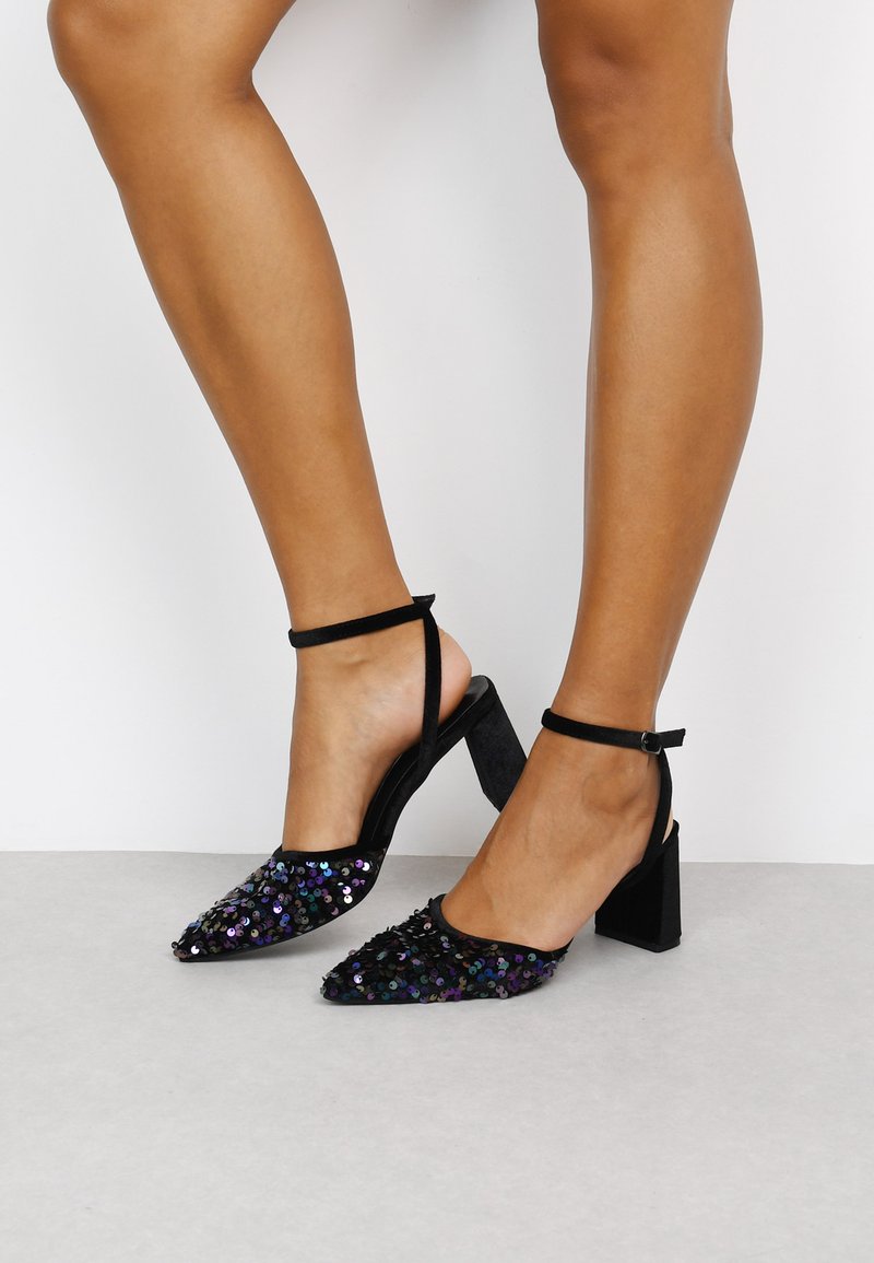 Zapatos de tacón negro con tira en el tobillo que presentan un tacón cuadrado, punta afilada y un upper cubierto de lentejuelas con acentos iridiscentes.