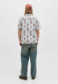 Chemise à manches courtes, bleu clair avec des motifs géométriques marron, associée à un jean bleu clair ample et des chaussures marron. Casquette rouge.