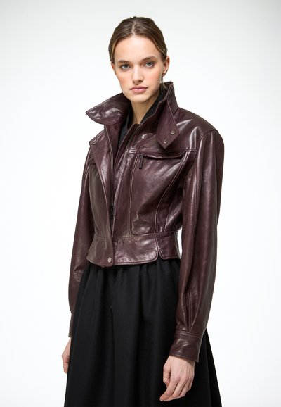 Femme portant une veste courte en cuir brun foncé avec un col montant et une fermeture éclair, associée à une jupe noire sur fond blanc.