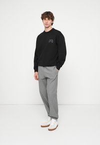 Μαύρο φούτερ με λογότυπο στο στήθος, γκρι φόρμα με ελαστικές μανσέτες και λευκά αθλητικά παπούτσια με ταμπά σόλες. Απλή, casual εμφάνιση.